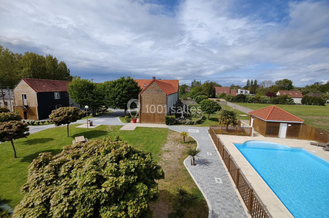 Vue d'un jardin avec pelouse, arbres, piscine entourée de dalles et maisons aux toits rouges sous un ciel partiellement…