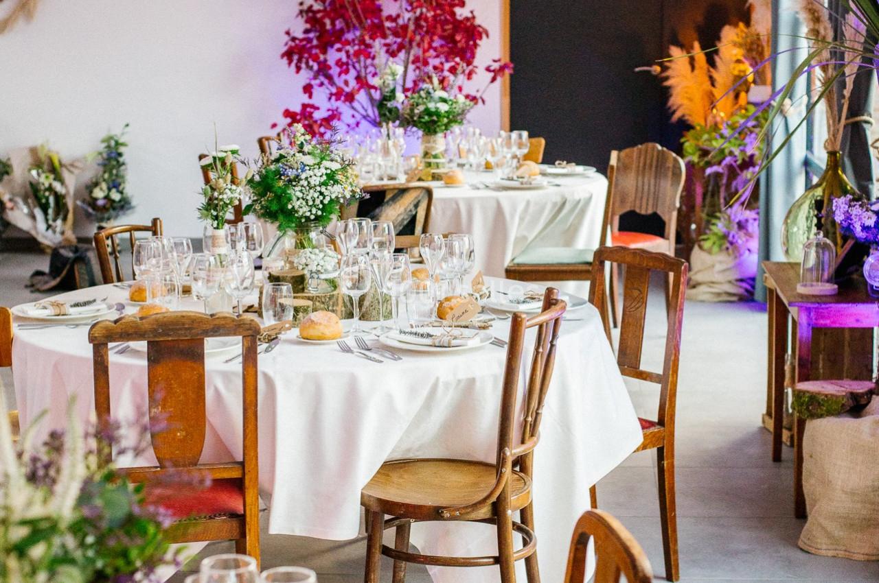 Salle décorée pour un événement avec tables rondes dressées, nappes blanches, vaisselle élégante et plantes en arrière-plan.