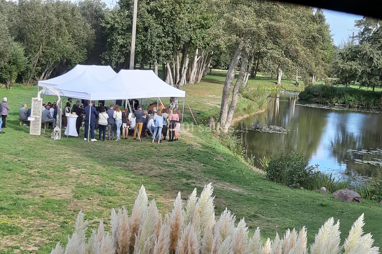 Groupe de personnes rassemblées sous une tente blanche près d'un étang entouré d'arbres et de verdure.