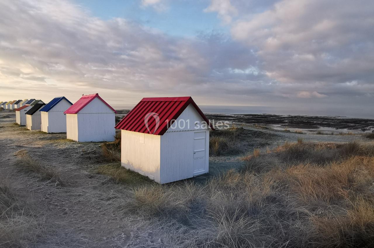 Location salle Gouville-sur-Mer (Manche) - Le Village du Phare #45 Alignement de petites cabanes blanches aux toits colorés sur une dune herbeuse, avec vue sur la mer et le ciel nuageux.