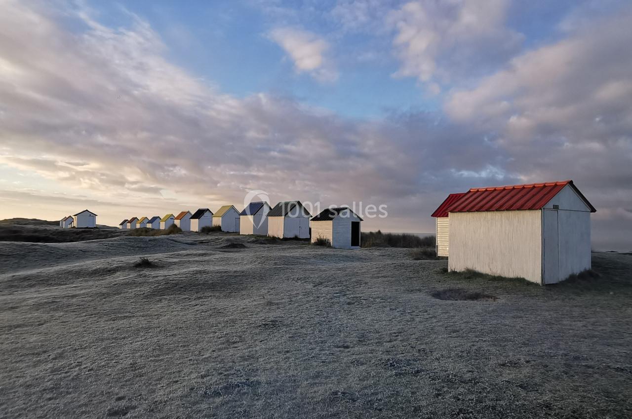 Location salle Gouville-sur-Mer (Manche) - Le Village du Phare #46 Alignement de petites cabanes blanches sur un terrain herbeux givré, sous un ciel nuageux au lever du jour.