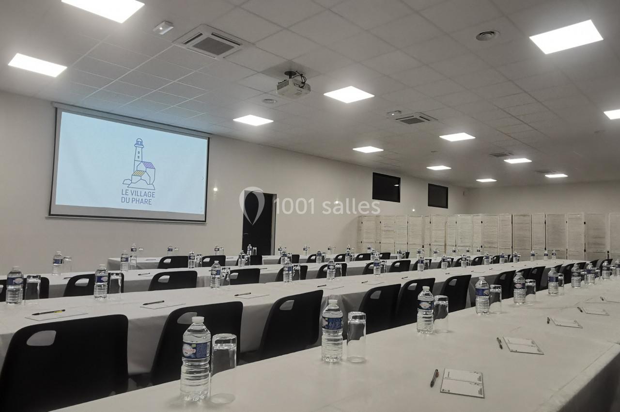 Location salle Gouville-sur-Mer (Manche) - Le Village du Phare #2 Salle de conférence avec des tables alignées, bouteilles d'eau, carnets, et un écran affichant un logo.