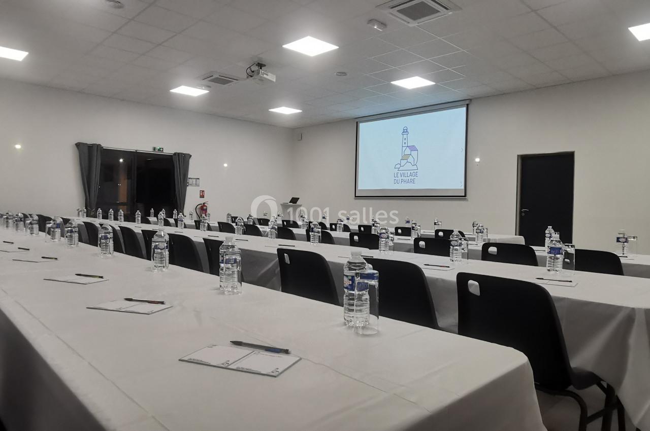 Location salle Gouville-sur-Mer (Manche) - Le Village du Phare #3 Salle de conférence aménagée avec des tables alignées, des bouteilles d'eau, des blocs-notes et un écran de projection…