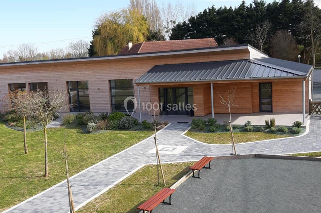 Location salle Gouville-sur-Mer (Manche) - Le Village du Phare #33 Bâtiment moderne en bois avec toit en métal, entouré d'un espace vert aménagé et de bancs sur une allée pavée.