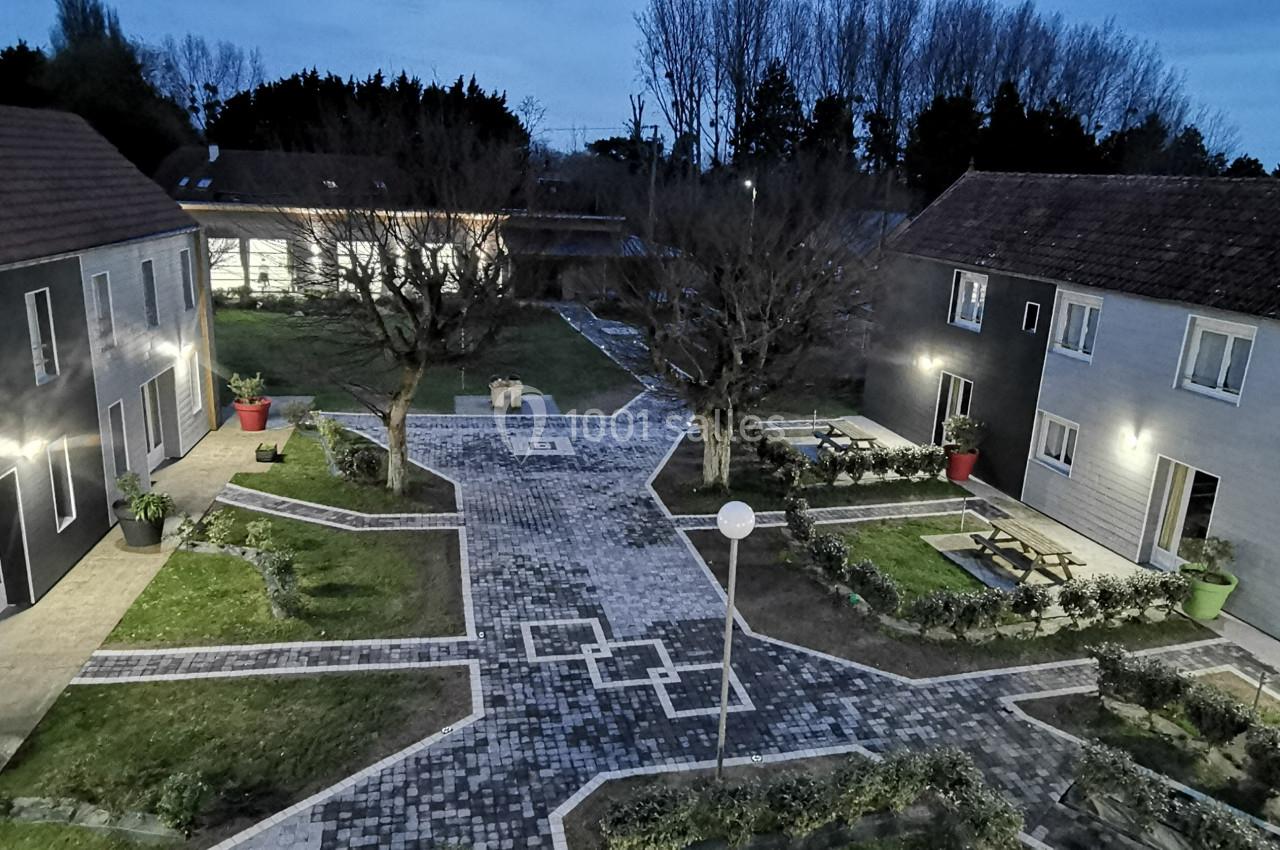 Location salle Gouville-sur-Mer (Manche) - Le Village du Phare #27 Cour pavée entourée de bâtiments modernes avec espaces verts, arbres et éclairage extérieur au crépuscule.