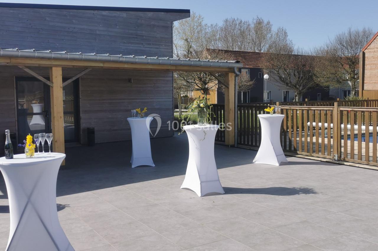 Location salle Gouville-sur-Mer (Manche) - Le Village du Phare #35 Tables hautes drapées de blanc disposées en extérieur sur une terrasse ensoleillée, avec quelques décorations florales.