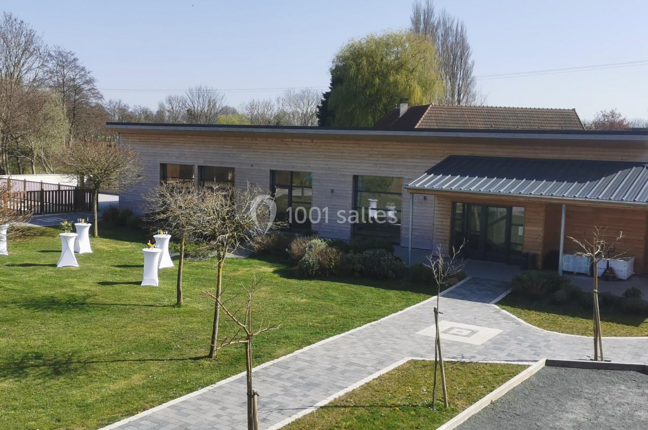 Location salle Gouville-sur-Mer (Manche) - Le Village du Phare #36 Bâtiment moderne en bois entouré d'un jardin avec des arbres et des tables hautes blanches en extérieur.