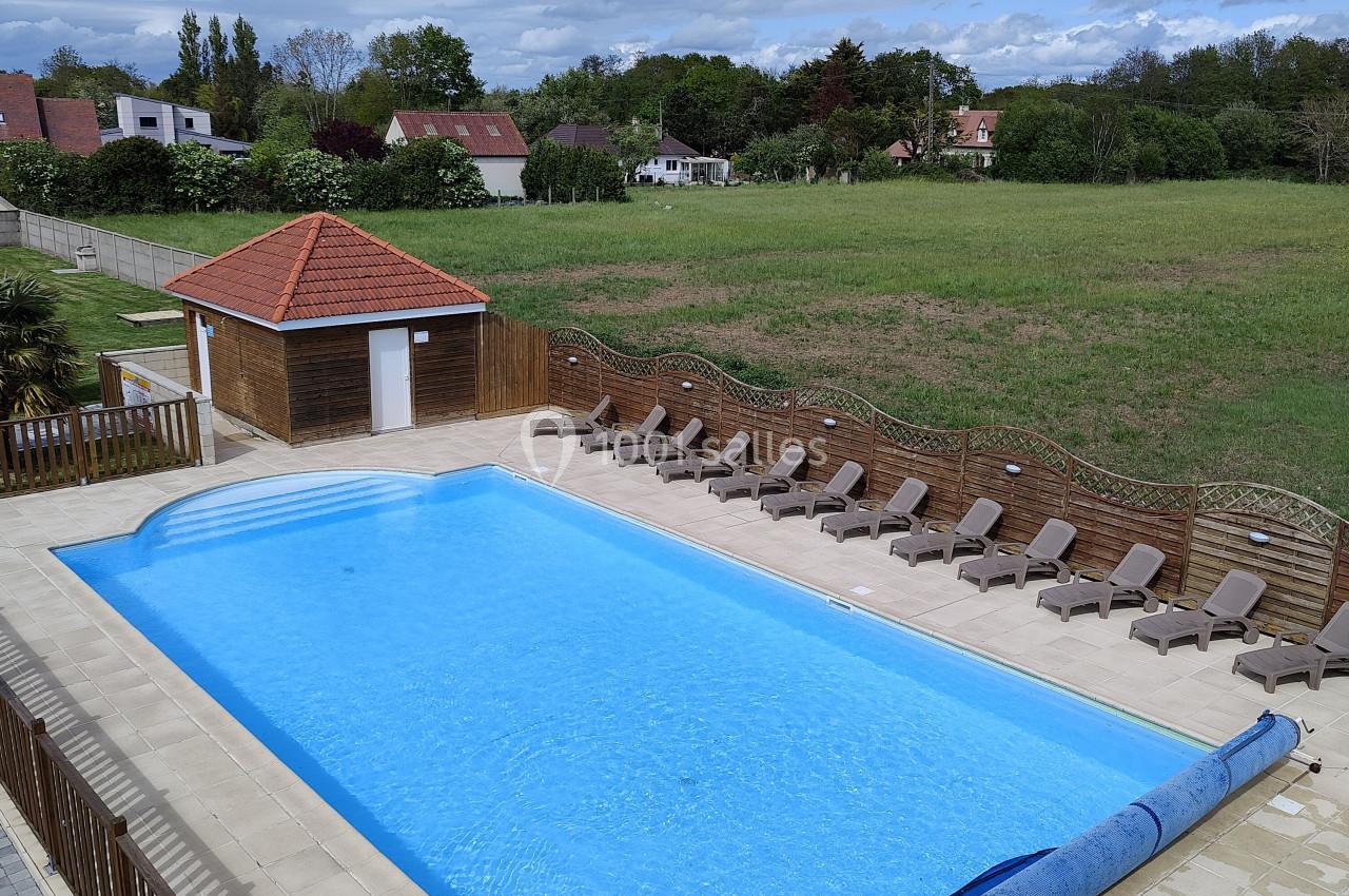Location salle Gouville-sur-Mer (Manche) - Le Village du Phare #28 Piscine extérieure rectangulaire entourée de transats, avec un abri en bois et un paysage verdoyant en arrière-plan.