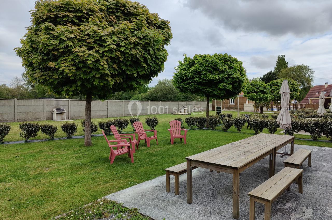 Location salle Gouville-sur-Mer (Manche) - Le Village du Phare #41 Table en bois avec bancs, chaises roses et parasol dans un jardin arboré, entouré de haies et clôtures grises.
