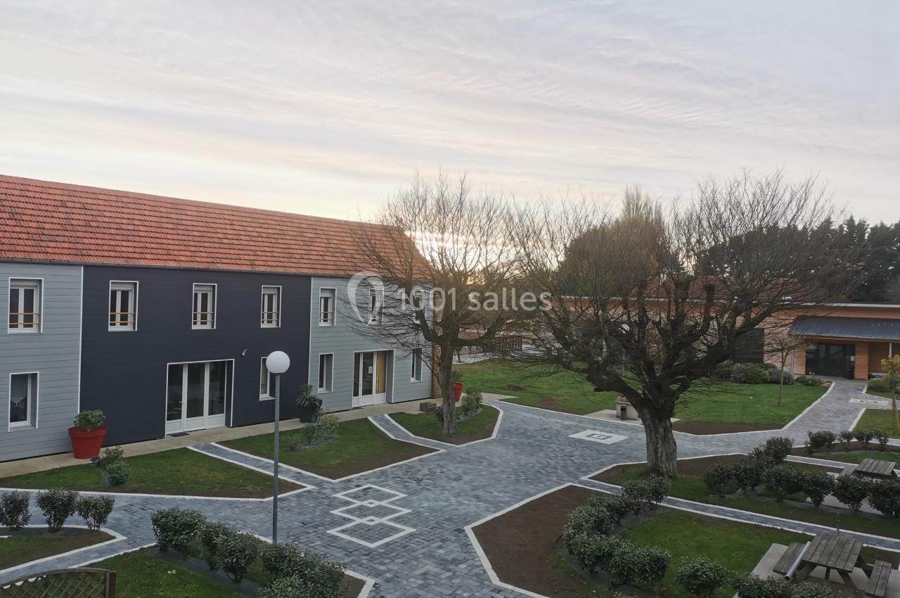 Location salle Gouville-sur-Mer (Manche) - Le Village du Phare #43 Vue d'une cour pavée avec bâtiments modernes, arbres taillés et bancs, sous un ciel partiellement nuageux.