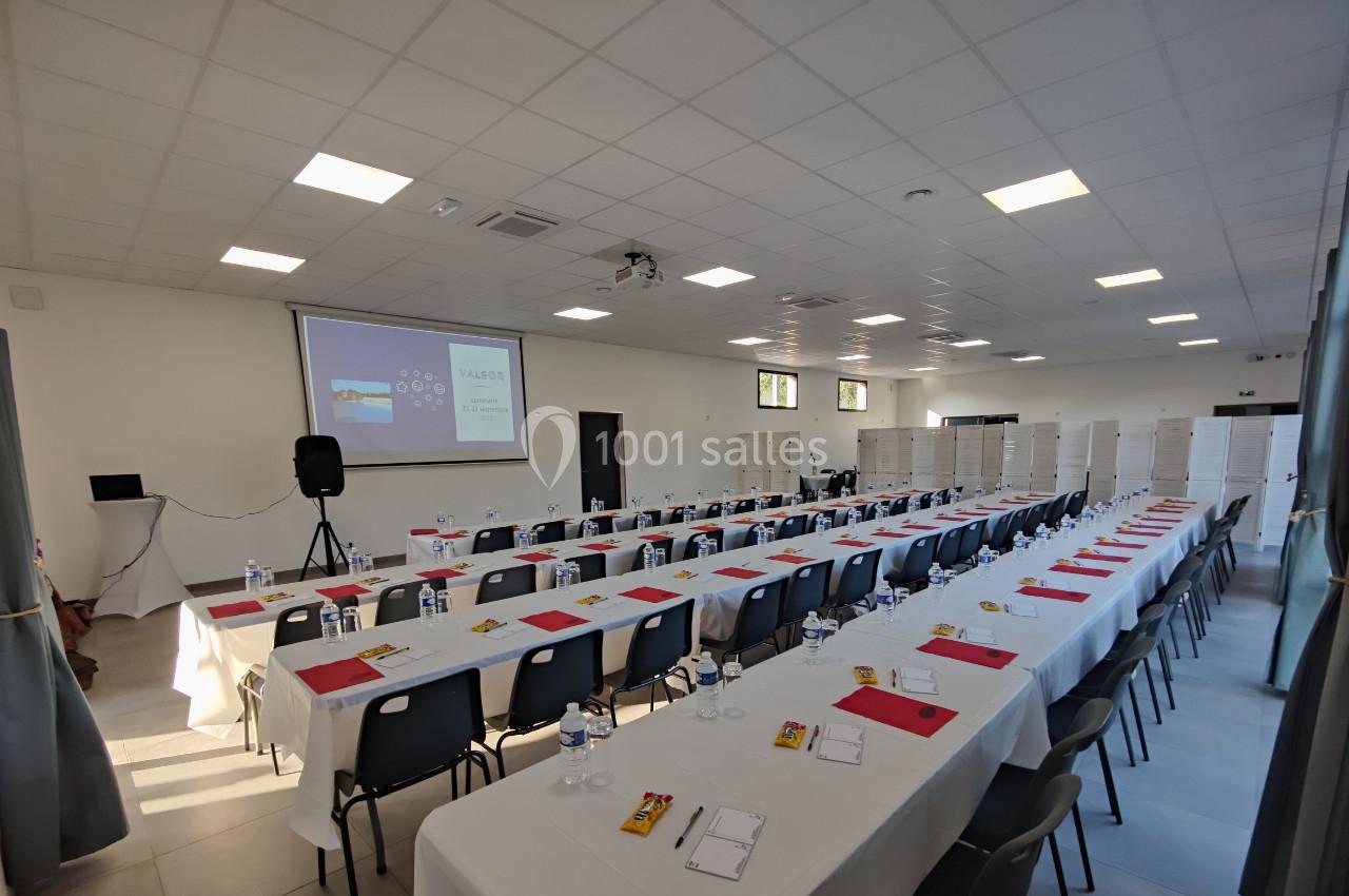 Location salle Gouville-sur-Mer (Manche) - Le Village du Phare #7 Salle de conférence avec des rangées de tables et chaises, écran de projection allumé et documents disposés sur les tables.
