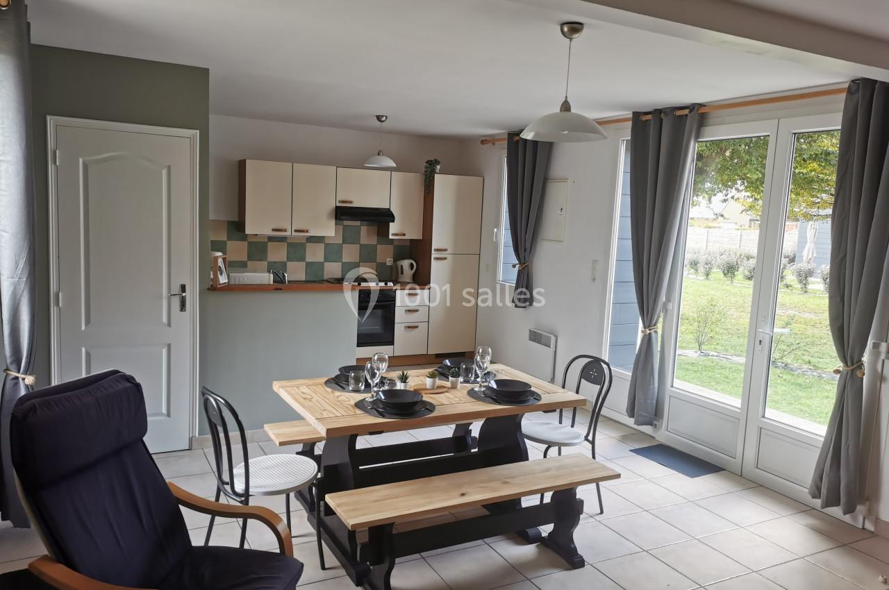 Location salle Gouville-sur-Mer (Manche) - Le Village du Phare #39 Salle à manger avec table en bois, chaises, cuisine ouverte et grandes fenêtres donnant sur un jardin.