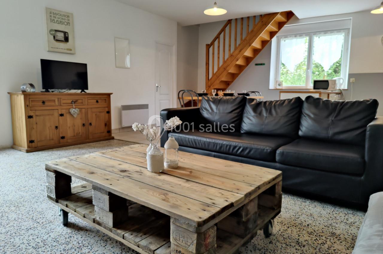 Location salle Gouville-sur-Mer (Manche) - Le Village du Phare #40 Salon lumineux avec canapé en cuir noir, table basse en palettes, meuble en bois, escalier et fenêtre donnant sur un jardin.