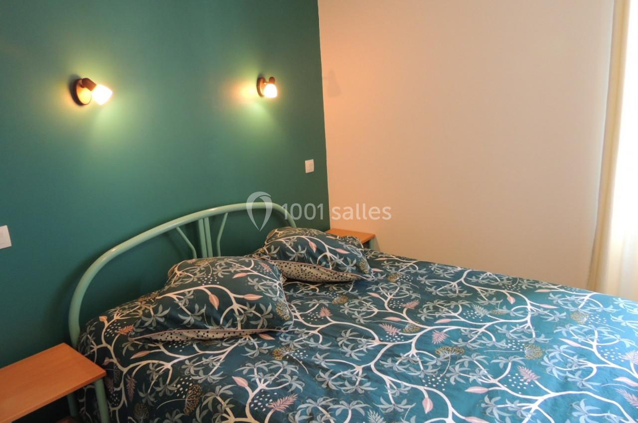 Location salle Gouville-sur-Mer (Manche) - Le Village du Phare #30 Lit double avec une tête de lit en métal vert, draps à motifs floraux, éclairé par deux appliques murales.