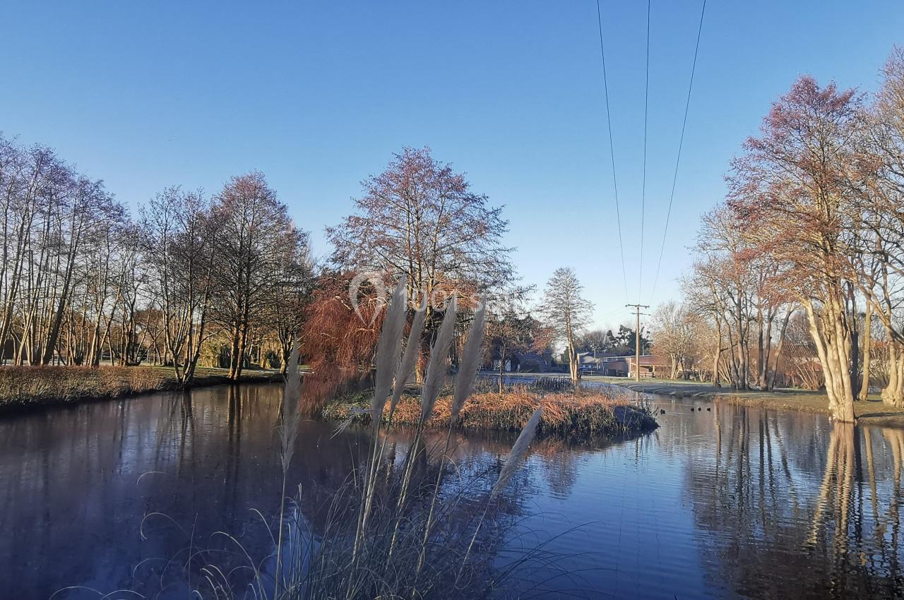 Location salle Gouville-sur-Mer (Manche) - Le Village du Phare #44 Plan d'eau entouré d'arbres aux couleurs automnales sous un ciel bleu, avec un petit pont visible au loin.