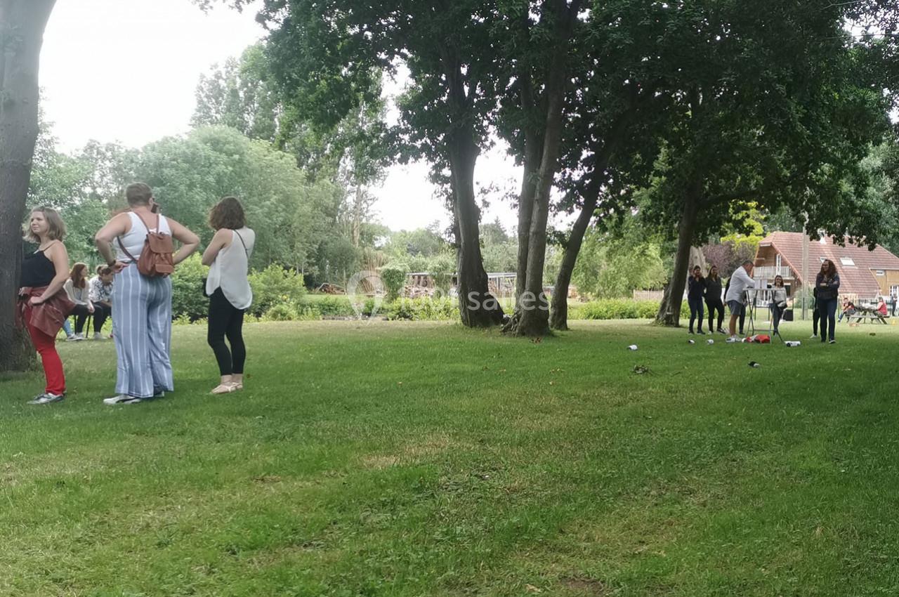 Location salle Gouville-sur-Mer (Manche) - Le Village du Phare #16 Personnes discutant ou jouant sur une pelouse entourée d'arbres, avec des bâtiments en arrière-plan.
