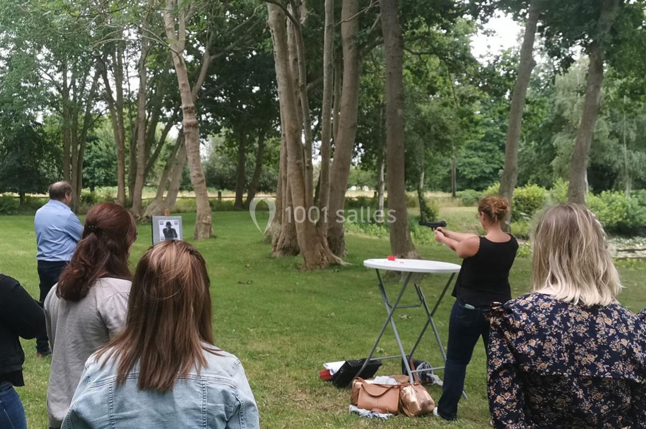 Location salle Gouville-sur-Mer (Manche) - Le Village du Phare #17 Une femme tire avec une arme à air comprimé sur une cible dans un parc, observée par plusieurs personnes.