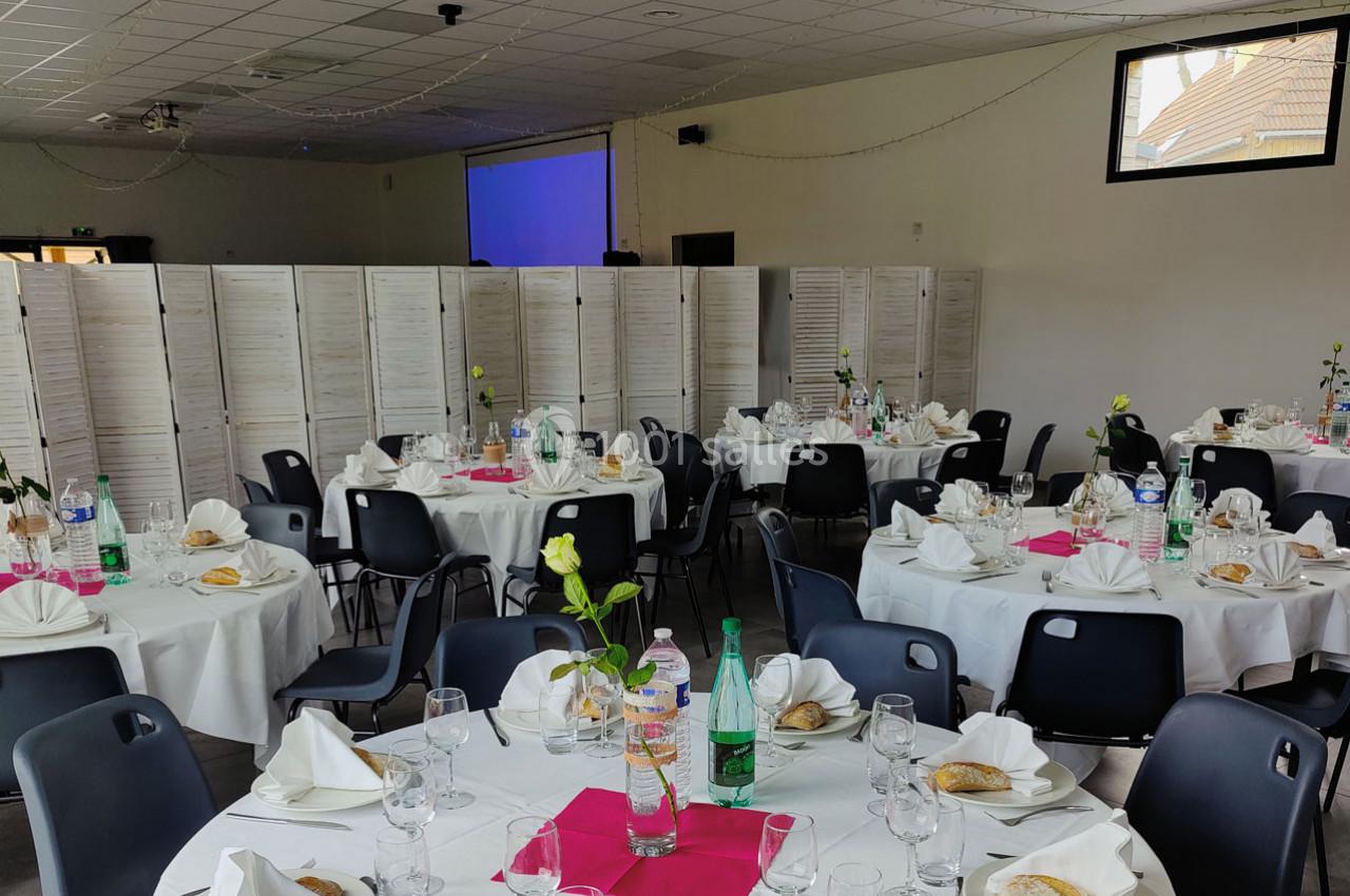 Location salle Gouville-sur-Mer (Manche) - Le Village du Phare #8 Salle de réception avec tables rondes dressées, nappes blanches, serviettes pliées, fleurs et bouteilles d'eau.