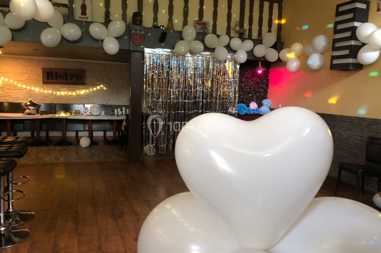 Salle décorée pour une fête avec des ballons blancs, guirlandes lumineuses et rideau argenté en arrière-plan.