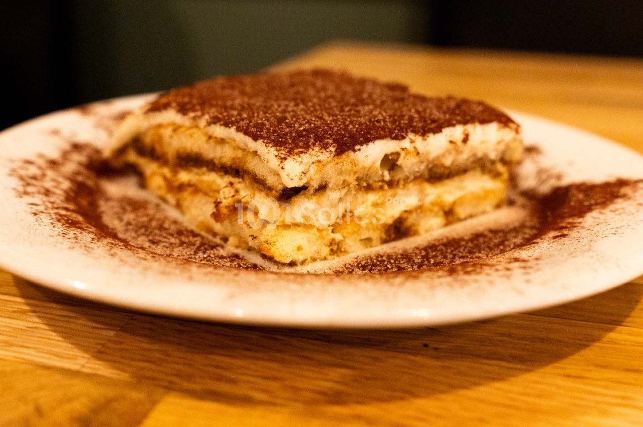 Part de tiramisu servie sur une assiette blanche, saupoudrée de cacao, posée sur une table en bois.