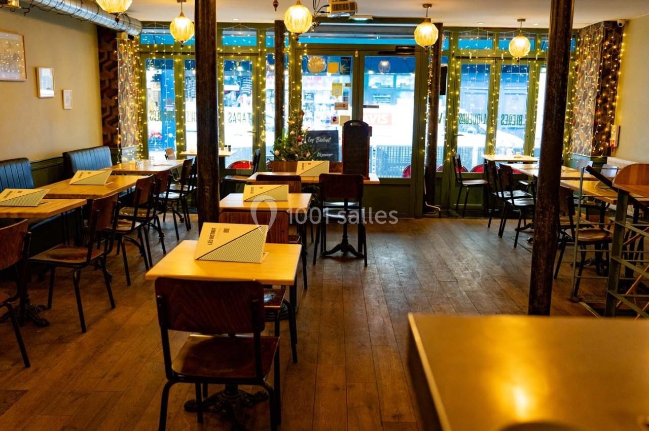 Salle de restaurant vide avec tables en bois, chaises, lumières suspendues et grandes fenêtres décorées de guirlandes…