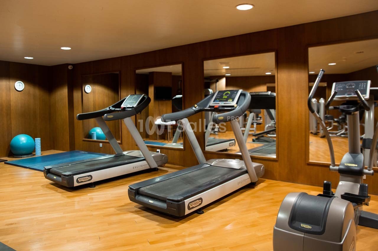Salle de sport avec tapis de course, vélo d'exercice, miroir mural et équipements de fitness sur parquet clair.