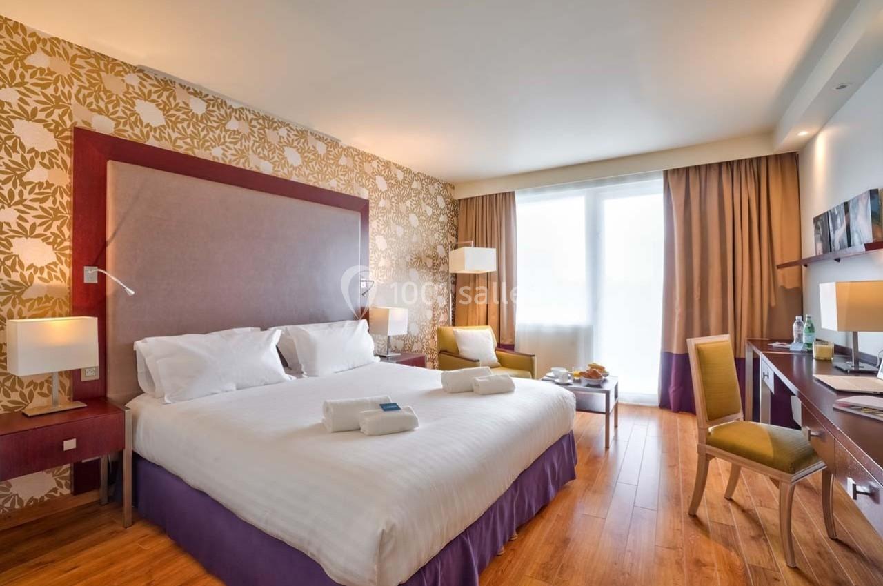 Chambre d'hôtel lumineuse avec lit double, bureau, fauteuils, parquet et grande fenêtre avec rideaux.