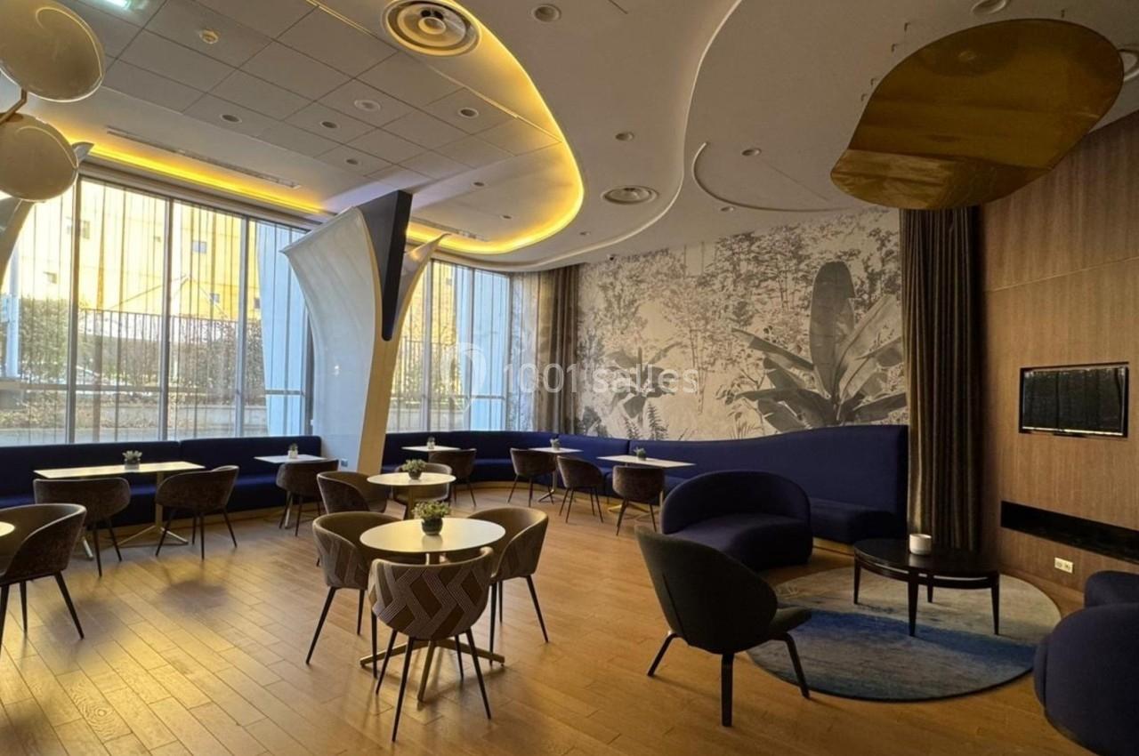 Salle moderne avec mobilier design, tables rondes, fauteuils bleus, grandes fenêtres et éclairage chaleureux.