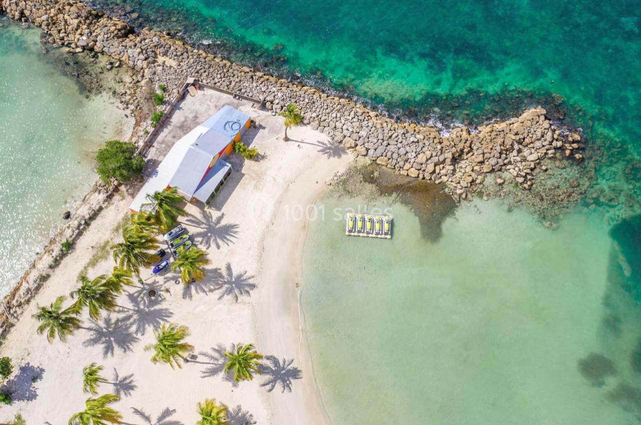Vue aérienne d'une plage tropicale avec palmiers, eaux turquoise, bâtiment blanc et embarcations alignées sur le sable.