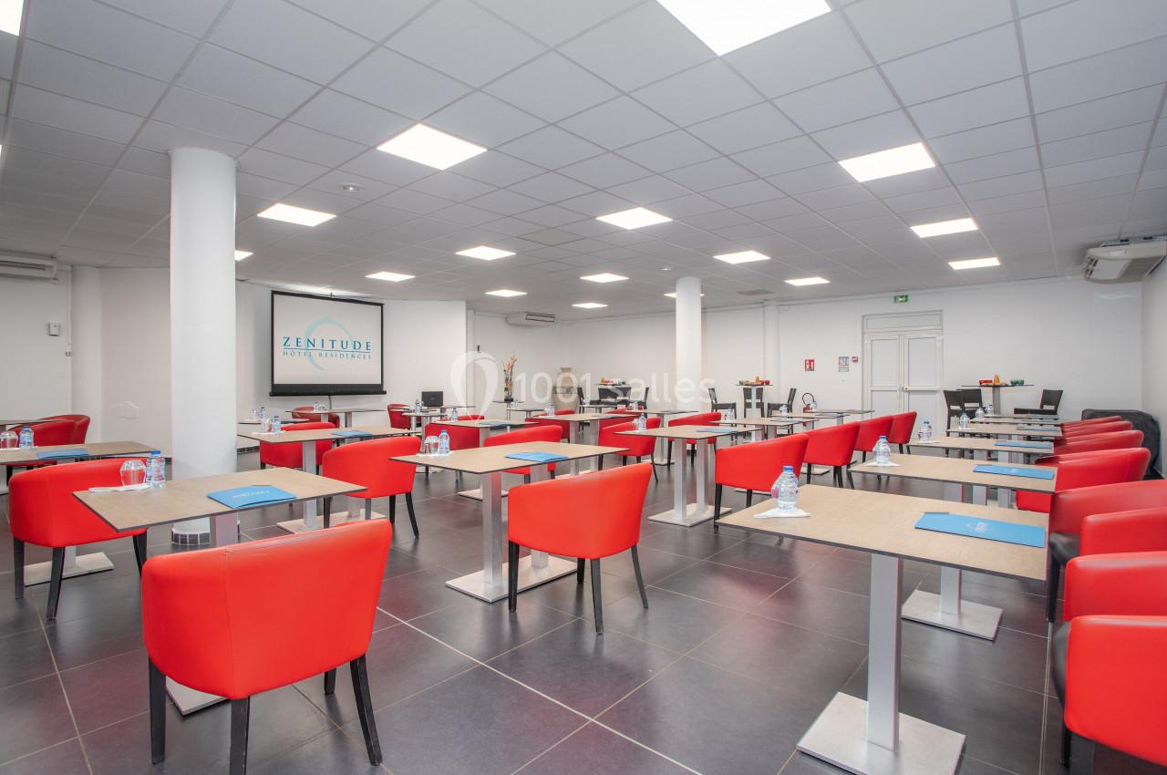 Salle de réunion lumineuse avec des tables individuelles, chaises rouges et matériel de conférence.