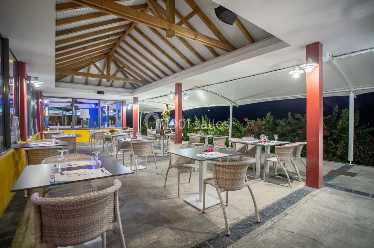 Salle de restaurant en terrasse couverte avec tables dressées, chaises en osier et vue sur un extérieur végétalisé.