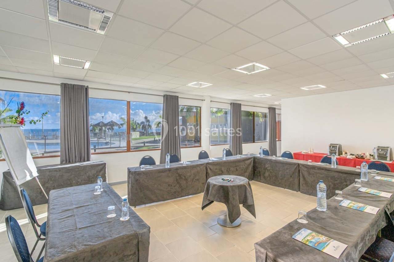 Salle de réunion lumineuse avec tables disposées en U, vue sur l'extérieur avec palmiers et mer à travers de grandes…