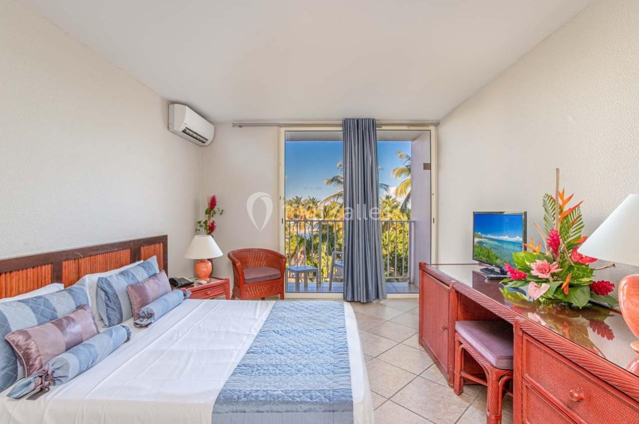 Chambre lumineuse avec lit double, mobilier en bois, télévision et balcon offrant une vue sur des palmiers.