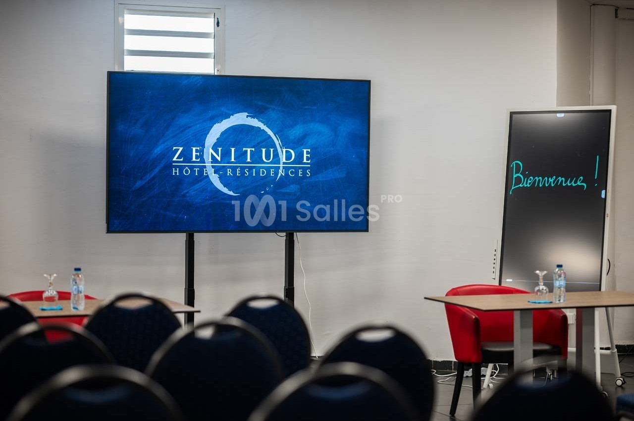 Salle de réunion avec écran affichant ’Zenitude Hôtel-Résidences’, tableau noir ’Bienvenue !’ et chaises disposées.