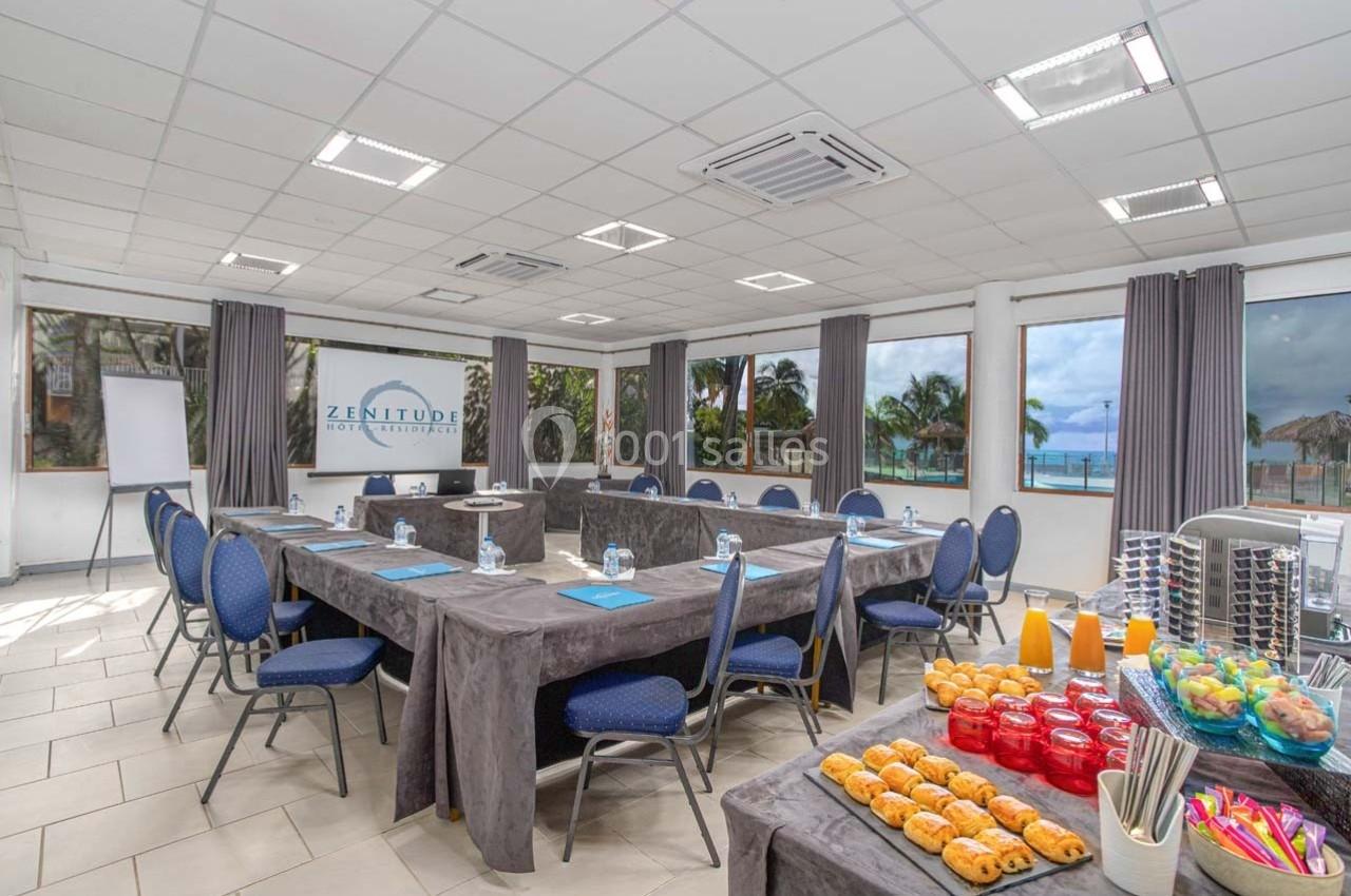 Salle de réunion lumineuse avec tables en U, chaises bleues, collations et vue sur la mer à travers de grandes fenêtres.