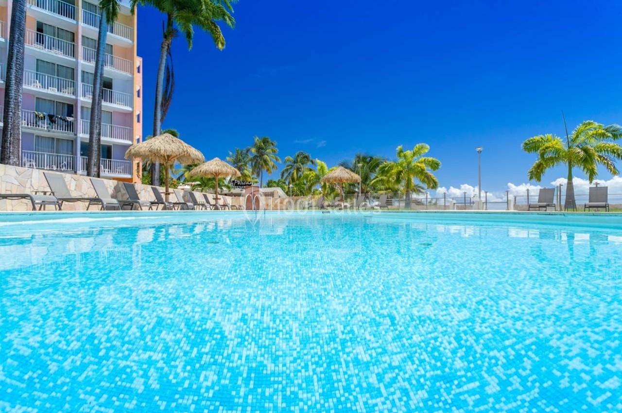 Piscine extérieure entourée de chaises longues, palmiers et parasols en paille sous un ciel bleu clair.
