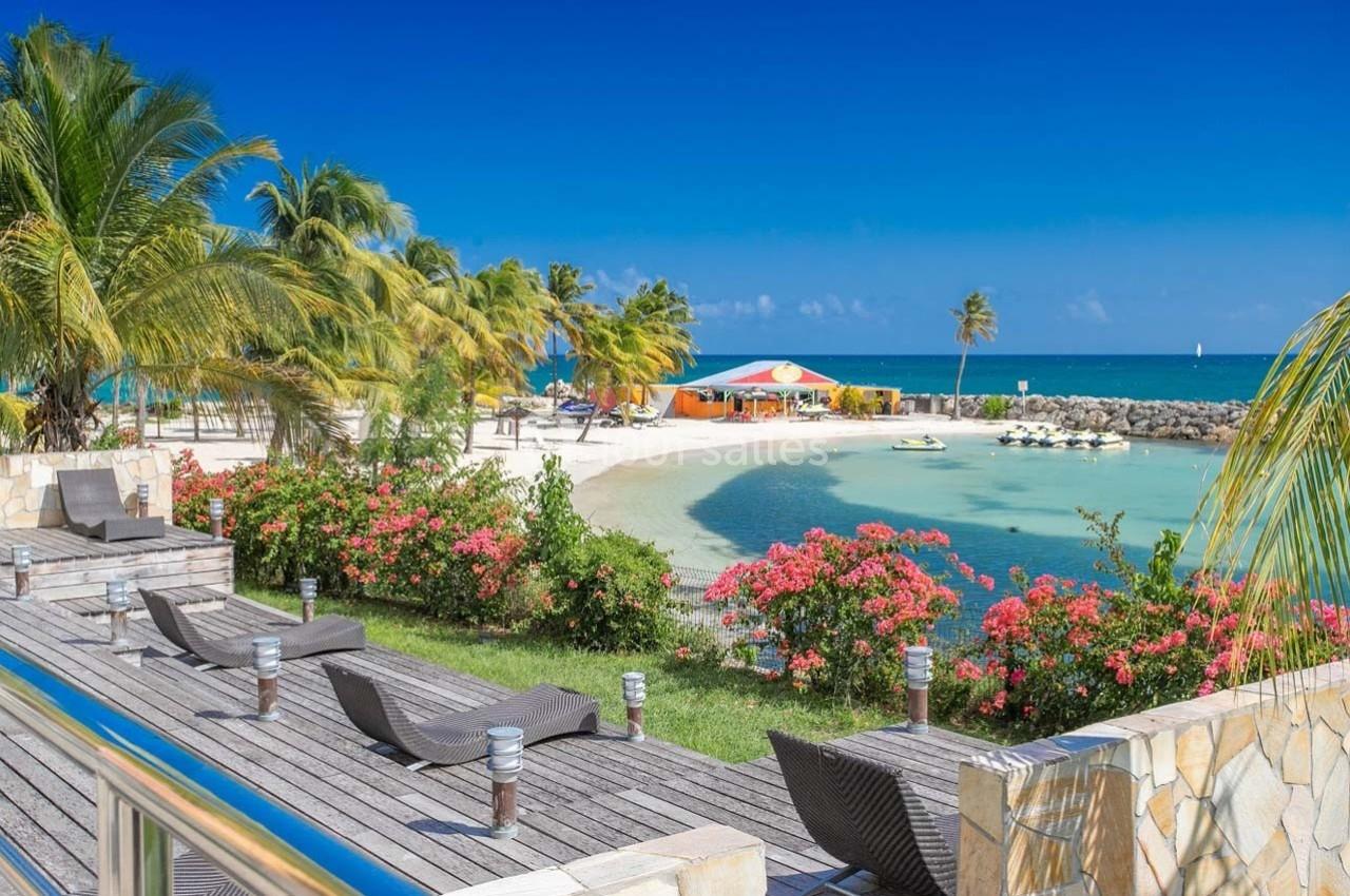 Plage tropicale avec palmiers, fleurs colorées, chaises longues, eau turquoise et un bâtiment coloré en arrière-plan.