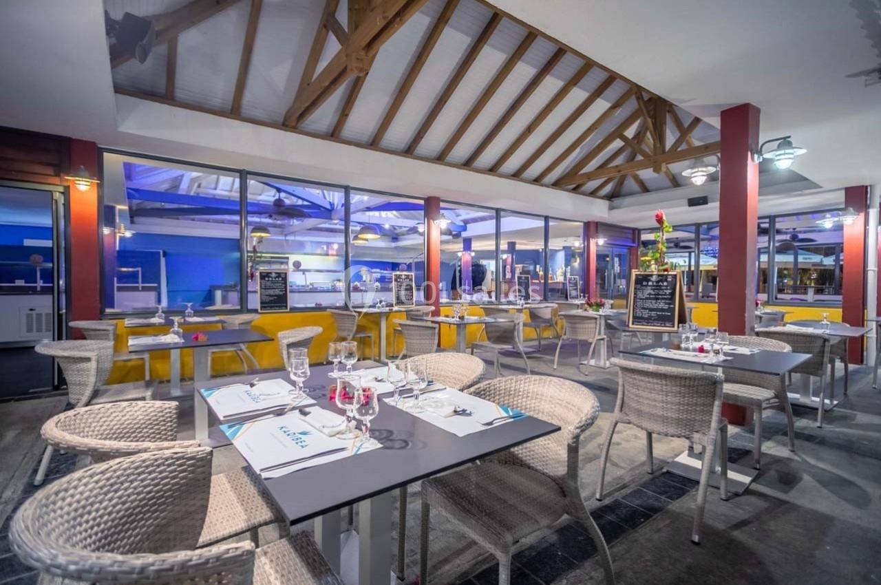 Salle de restaurant lumineuse avec tables dressées, chaises en osier et vue sur un comptoir ouvert en arrière-plan.