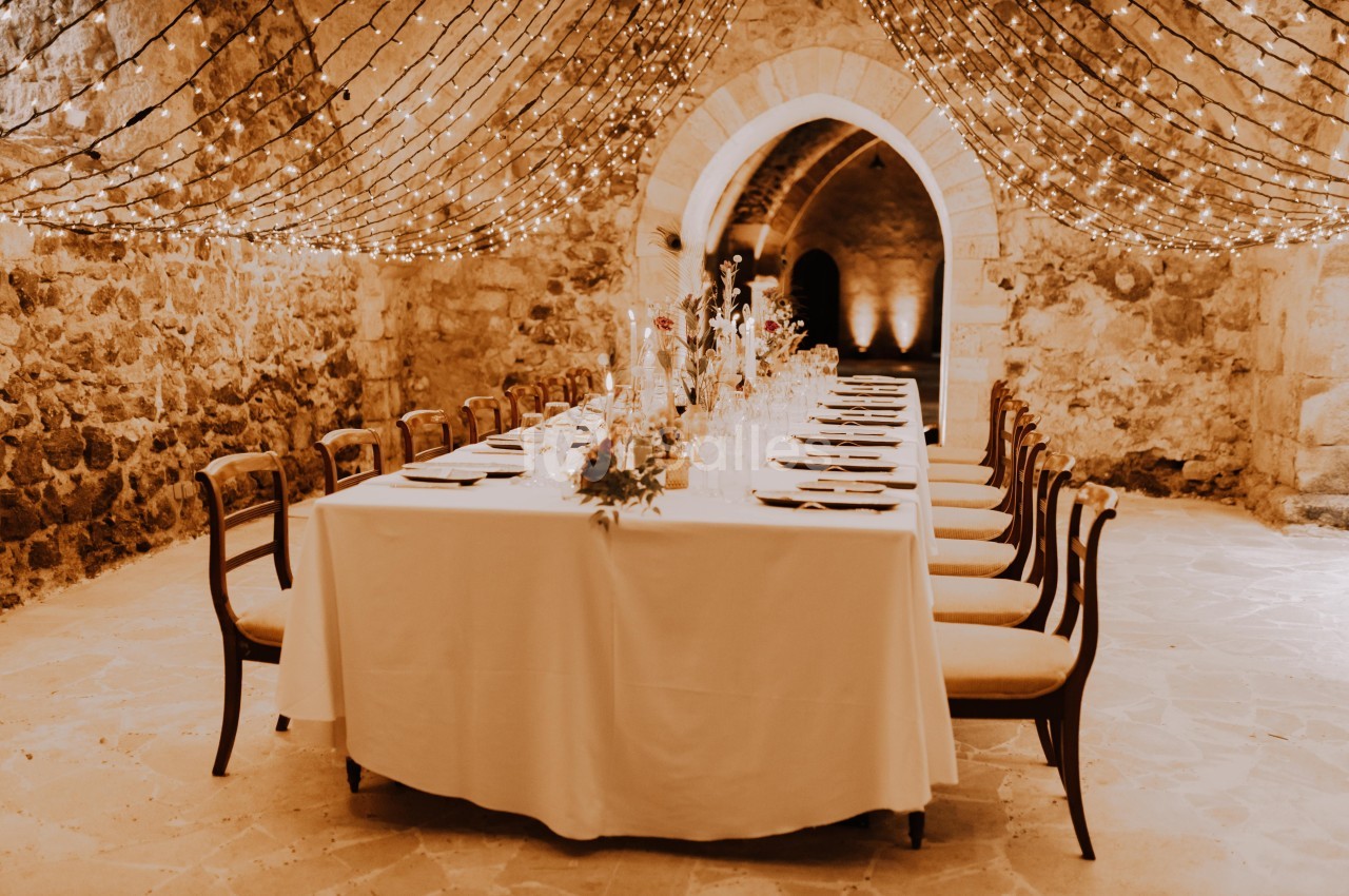 Table dressée pour un dîner dans une salle voûtée en pierre, éclairée par des guirlandes lumineuses suspendues.