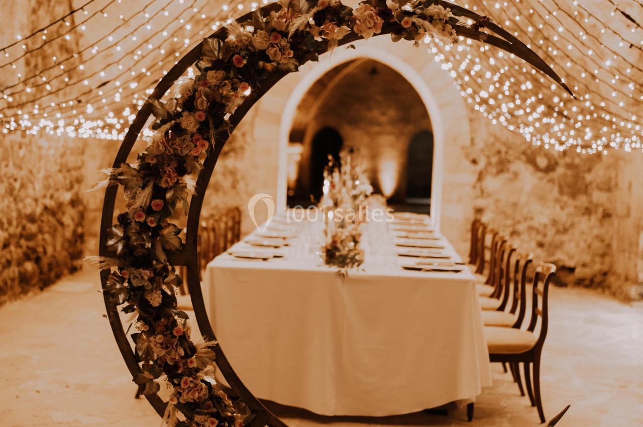 Décoration de mariage avec une arche en forme de lune ornée de fleurs, entourée de bougies, sous des guirlandes lumineuses.