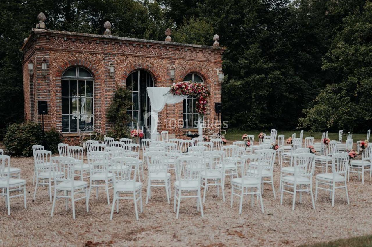 Chaises blanches disposées en extérieur devant une petite bâtisse en briques décorée pour une cérémonie.