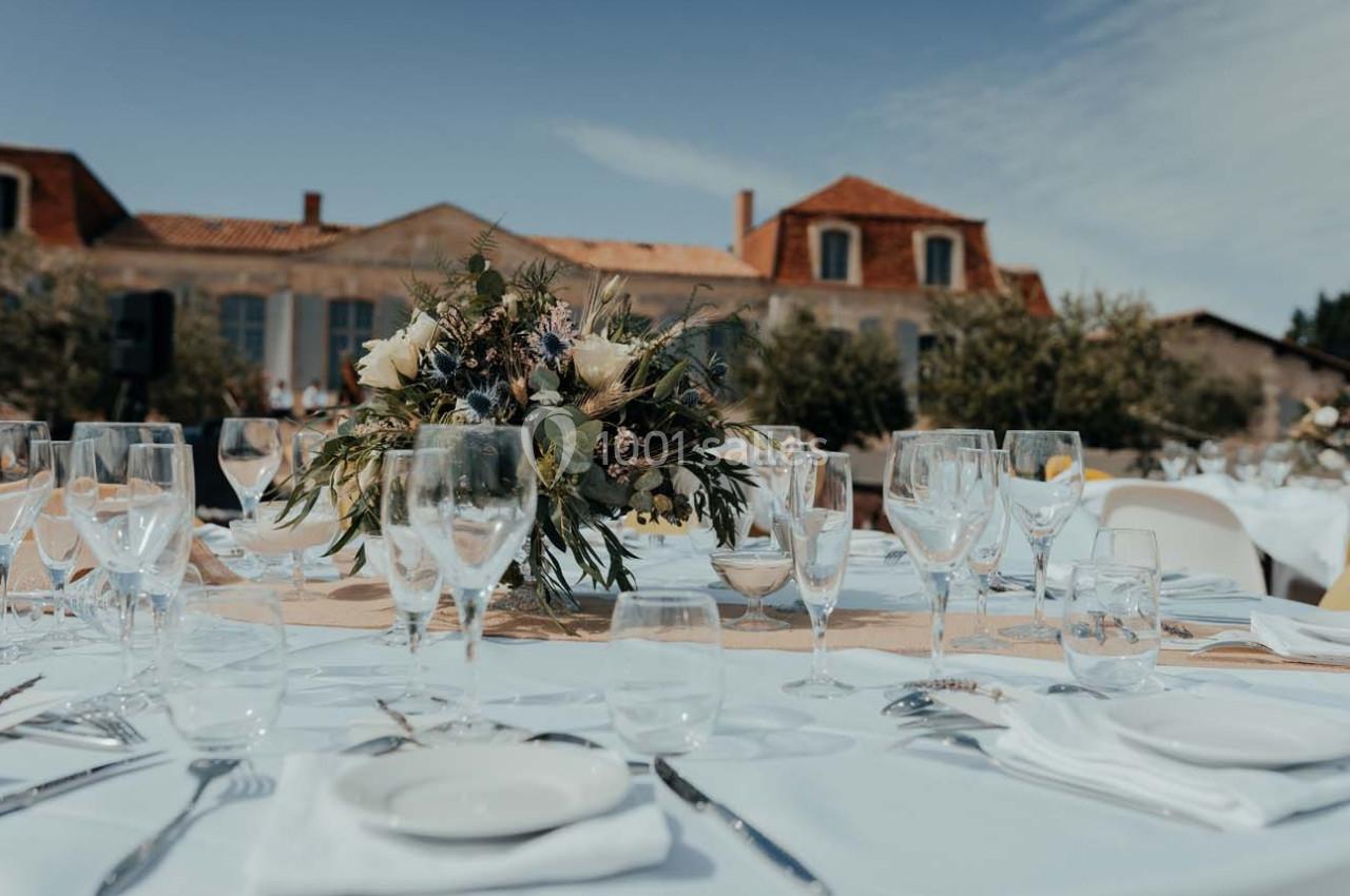 Table dressée avec nappes blanches, verres et centre de table floral, en extérieur devant un bâtiment en pierre.