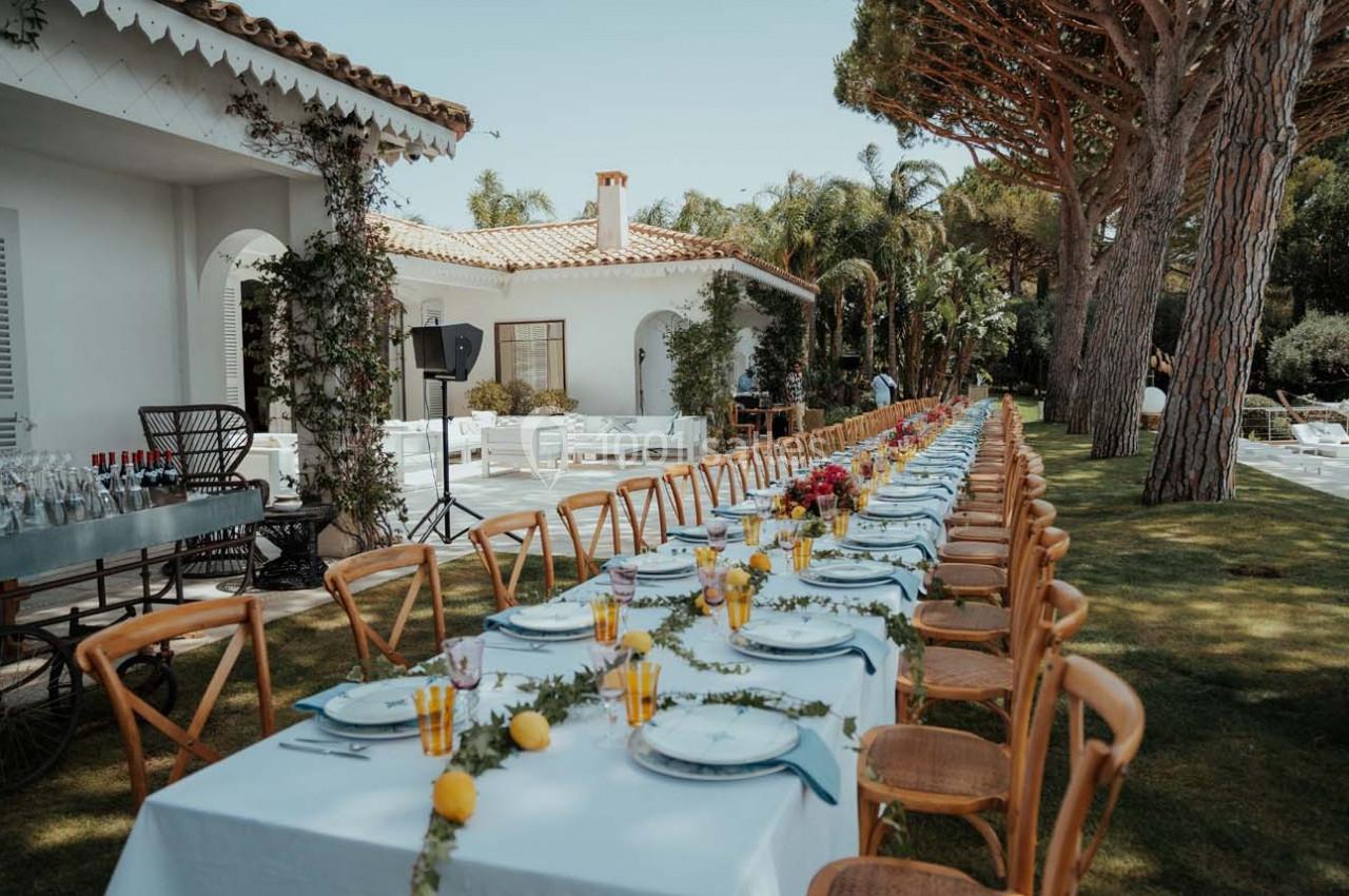 Grande table dressée en extérieur avec vaisselle, décorations florales et citrons, entourée de chaises en bois.