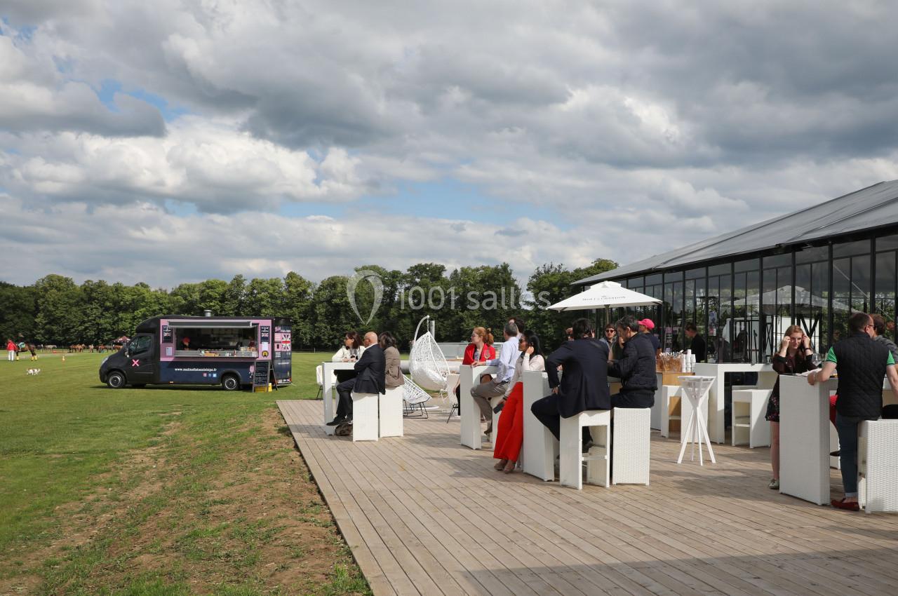 Personnes assises à des tables en extérieur près d'une verrière, avec un food truck et une pelouse en arrière-plan.