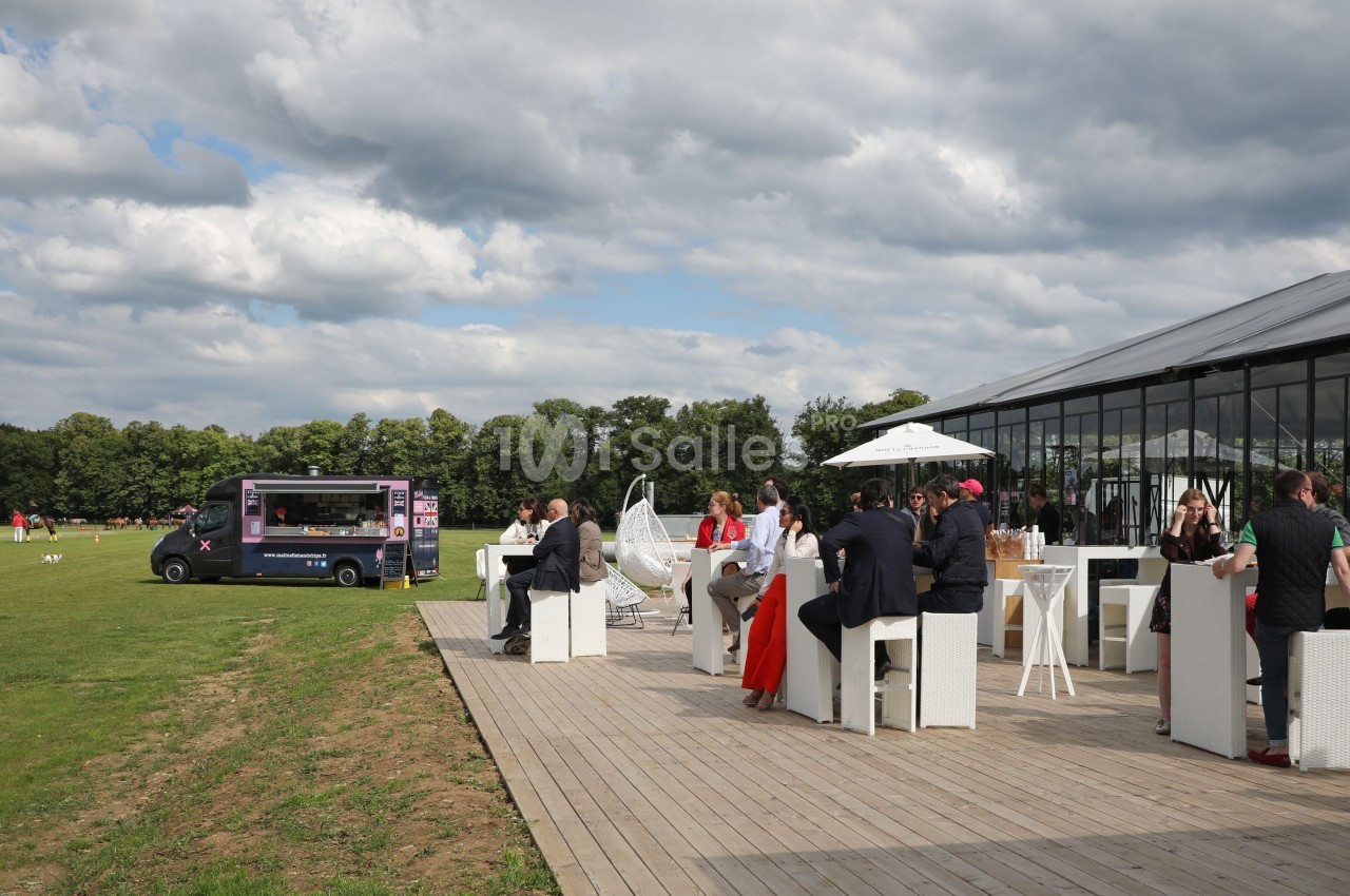 Personnes assises à des tables en extérieur près d'une verrière, avec un food truck et une pelouse en arrière-plan.