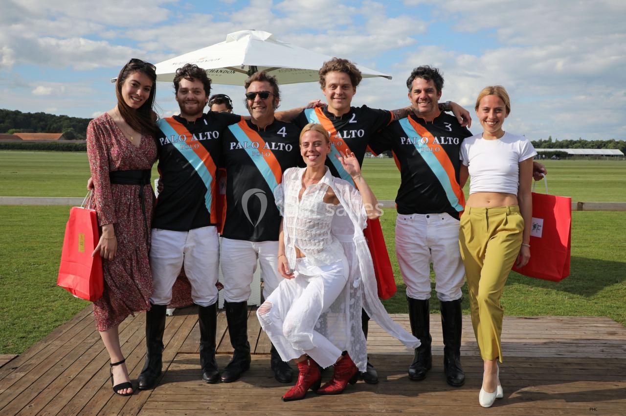 Un groupe de personnes pose souriantes sur une terrasse en bois, avec des joueurs de polo en tenue au centre.