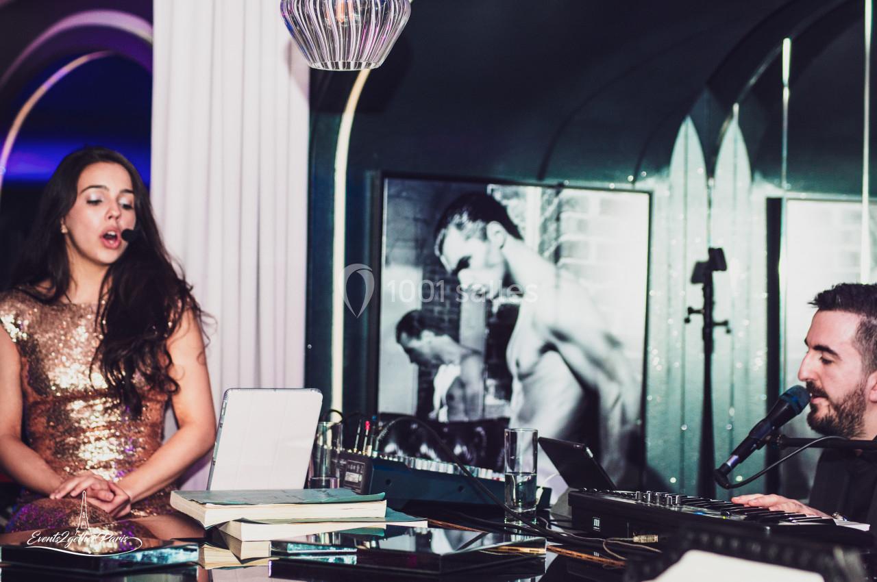 Une chanteuse en robe dorée et un musicien jouent dans un intérieur élégant avec des photos en arrière-plan.