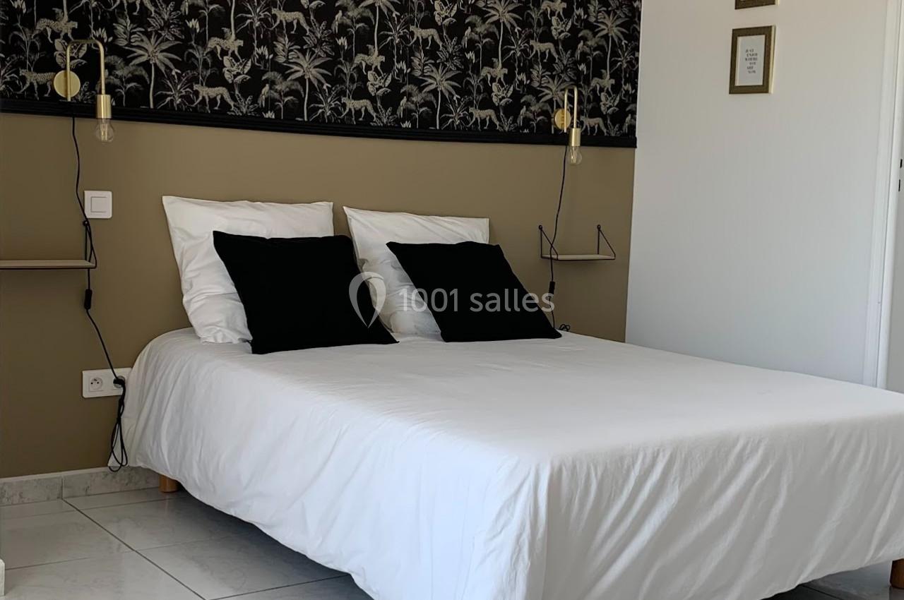 Lit double avec draps blancs, coussins noirs, éclairage mural et décoration murale à motifs végétaux.