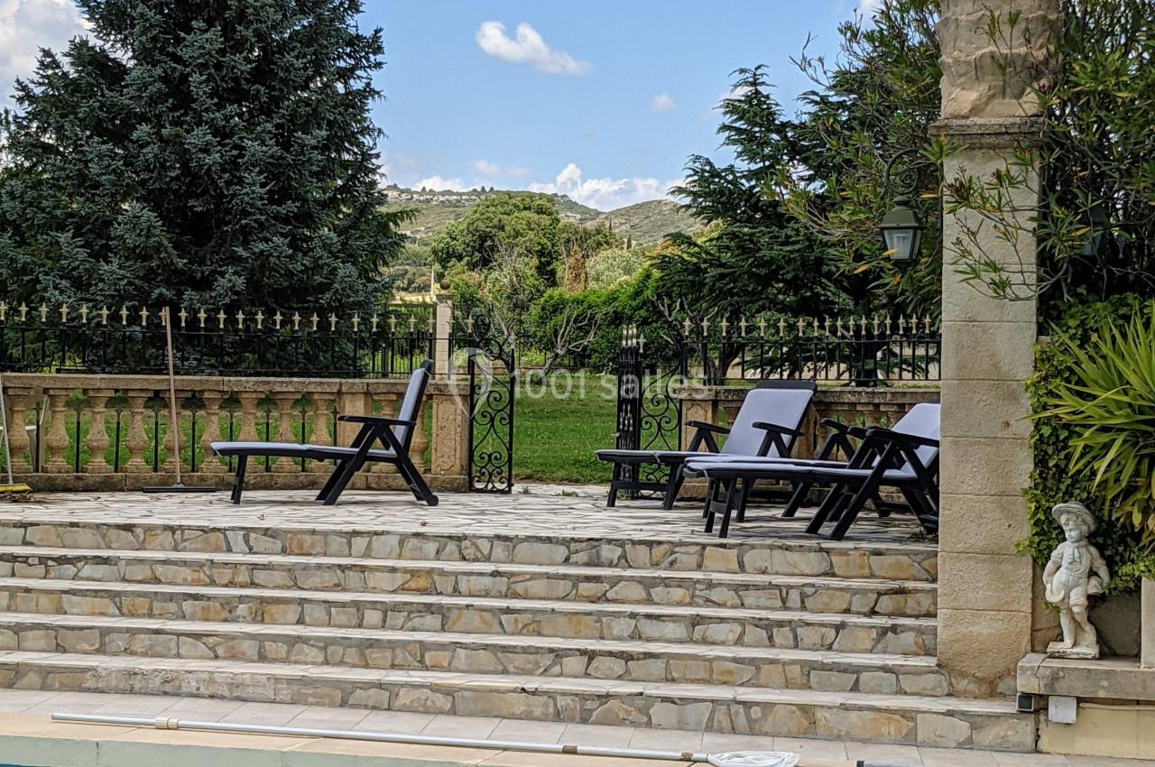 Terrasse en pierre avec chaises longues près d'une piscine, entourée de verdure et d'arbres sous un ciel bleu.