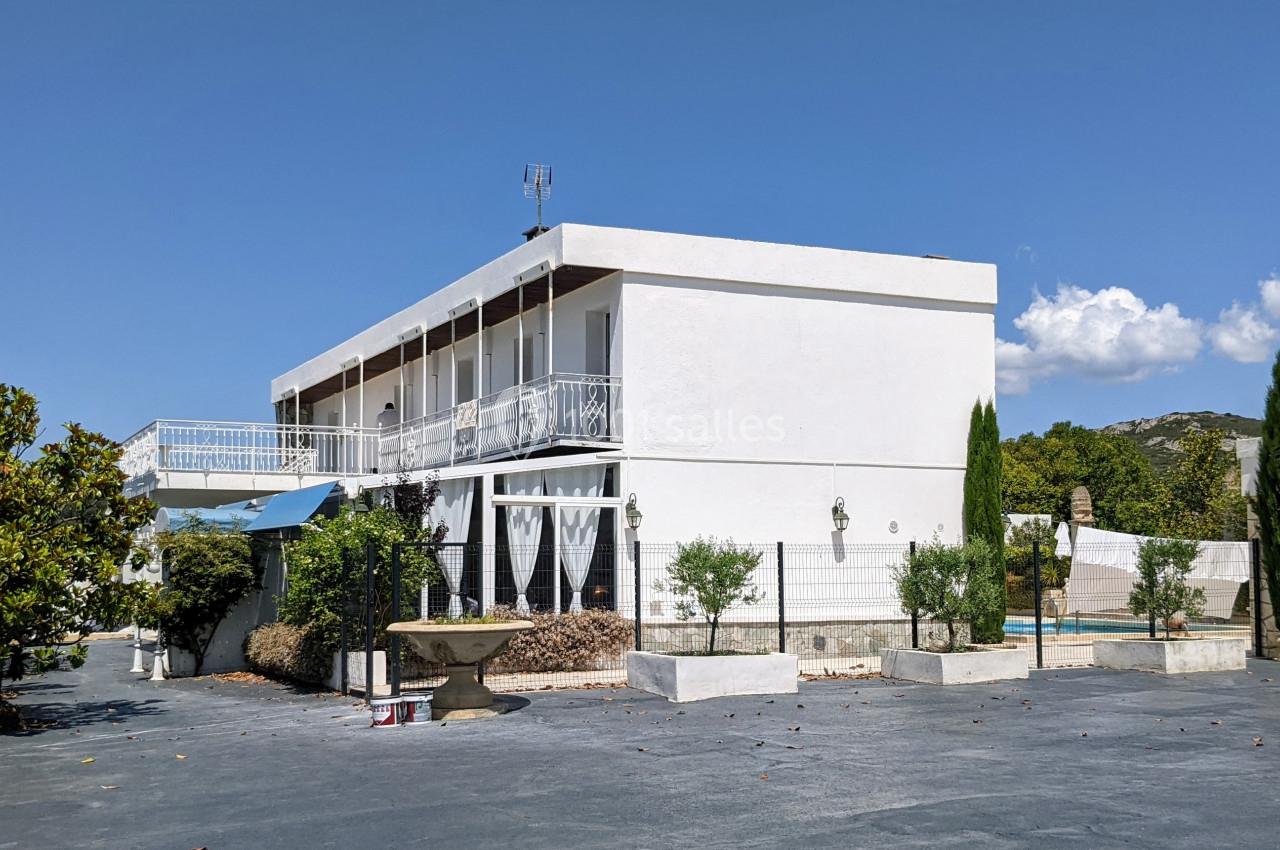 Maison blanche à deux étages avec balcon, entourée d'arbres et d'une cour asphaltée sous un ciel bleu.